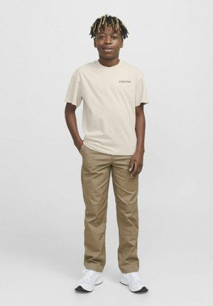 Camiseta de algodón beige con "Club1990" impreso en negro, combinada con pantalones marrones y zapatillas blancas. El modelo está de pie con las manos en los bolsillos.
