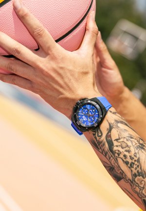 Montre avec un bracelet en caoutchouc bleu, boîtier rond noir, cadran bleu et plusieurs fonctionnalités de chronographe. Main tenant un ballon de basket rose.