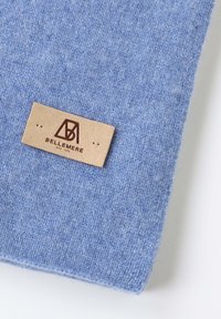 Écharpe en laine bleue avec une texture douce. Étiquette en cuir attachée avec un logo embossé et le nom de la marque, positionnée sur le bord inférieur.