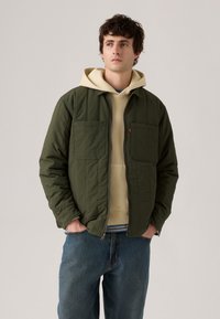 Veste en olive matelassée avec deux poches avant, associée à un sweat à capuche beige. Portée avec un jean en denim bleu clair. Détails de couture subtils.