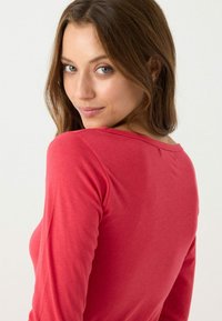 Camisa ceñida de manga larga en color coral intenso, confeccionada en mezcla suave de algodón con escote redondeado y detalles mínimos en las costuras de los hombros.