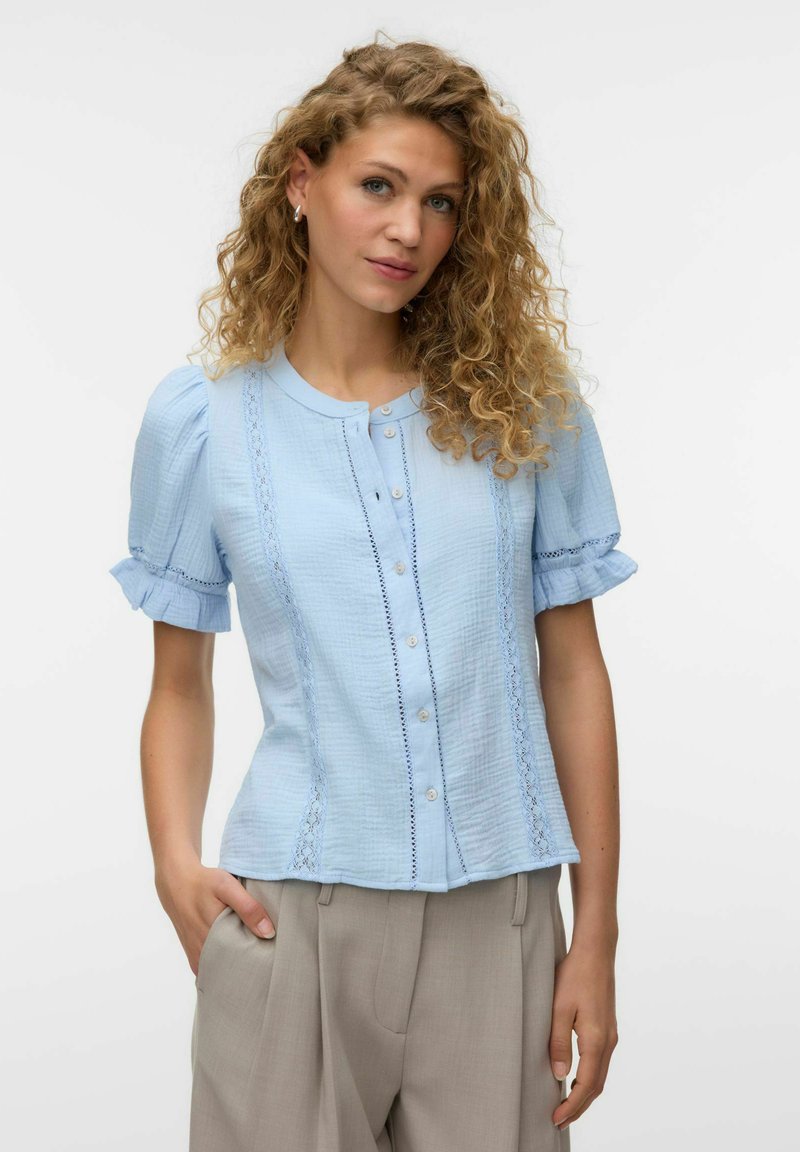Femme aux cheveux bouclés portant un chemisier bleu clair boutonné avec des manches bouffantes et un pantalon beige taille haute, posant avec une main dans la poche.
