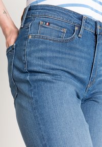 Modré džínové kalhoty s úzkým střihem, se světlými švy a malou červenou, bílou a modrou nášivkou na přední kapse.