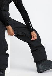 Zwarte skibroek met een ritsopening aan de zijkant, verstelbare taille en elastische enkels. Kenmerken zijn onder andere subtiele branding op de mouw.