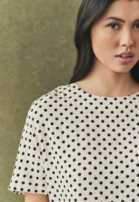 Cremefarvet bluse med sorte prikker, rund halsudskæring og korte ærmer. Stoffet har en blød tekstur og en løs pasform.