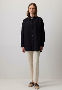 Una donna si trova davanti a uno sfondo semplice, indossando una camicia nera oversize con bottoni, pantaloni dritti color avorio e scarpe beige con cinturini neri.