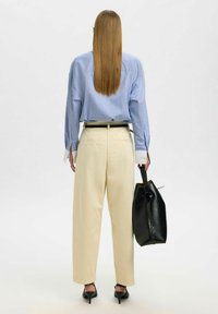 Chemise oversize rayée bleu clair, pantalon crème et sac à main noir. Talons hauts et cheveux longs visibles de dos. Design simple et moderne.