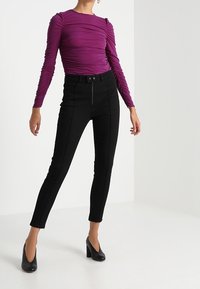 Figurbetontes Langarmshirt aus strukturiertem lila Stoff, kombiniert mit hoch taillierten schwarzen Skinny-Pants mit Reißverschluss vorne und Nahtdetails. Schwarze High Heels.