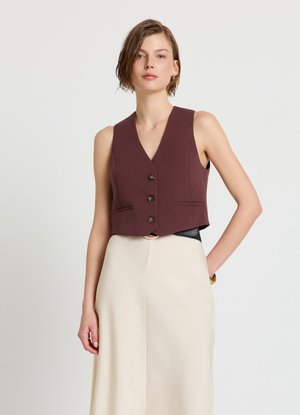 Femme aux cheveux courts châtains portant un gilet sans manches marron foncé boutonné et un pantalon beige à jambes larges et taille haute, debout devant un fond uni.