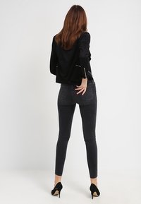 Jean noir ajusté taille haute, poches arrière détaillées et légère brillance, associé à une veste noire et des escarpins noirs.