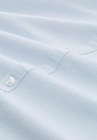 Țesătură de cămașă albastră deschisă, cu o textură netedă, având detalii cu buton și cusături subtile. Suprafața este ușor șifonată.