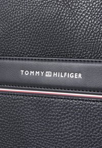 Cuero negro texturizado con una banda horizontal lisa que presenta el texto "TOMMY HILFIGER" en blanco, acentuado por una franja roja y blanca.