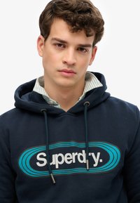 Granatowy hoodie z troczkami, z wyraźnym logo "Superdry" w bieli i turkusie, noszony na kolorowej koszuli w paski.
