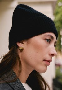 Bonnet en maille côtelée noire avec un sommet pointu ; boucle d'oreille en or en forme d'anneau ; modèle à la peau claire et aux cheveux bruns. Accent sur la texture et les matériaux.