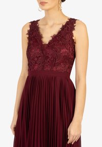 Robe de soirée bourgogne avec un corsage en dentelle, jupe plissée et décolleté en V. Présente des broderies florales élaborées et une taille cintrée.