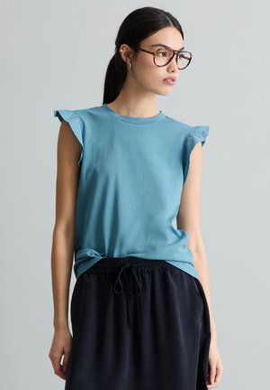 Frau mit dunklem Haar und Brille, die ein blaues ärmelloses Oberteil mit Rüschen an den Schultern und schwarze Hosen mit Kordelzug trägt.