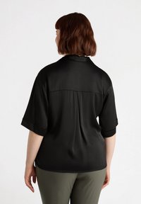 Blusa negra de manga corta con textura suave, detalle de yugo en la parte posterior y puños doblados. Combinada con pantalones verde caqui.