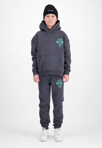 Jongen met een donkergrijze hoodie en bijpassende joggingbroek met turquoise berenlogo's, zwarte gebreide muts en witte sneakers, staand tegen een witte achtergrond.