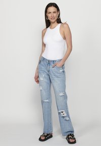 Bela bodysuit brez rokavov, svetlo modri raztrgani džins, masivni črni sandali s srebrnimi detajli, model pozira z eno roko v žepu.