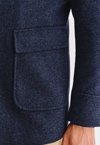 Marynarski wełniany płaszcz z dużą kieszenią naszywaną po lewej stronie. Tkanina ma teksturowane wykończenie, dopasowany krój i usztywniane rękawy.