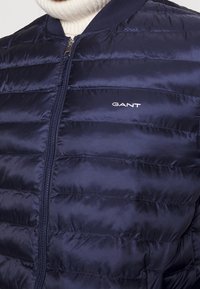 Giubbotto imbottito blu navy con motivi orizzontali cuciti, chiusura con zip e colletto a costine. Presenta il logo GANT in bianco sul petto.
