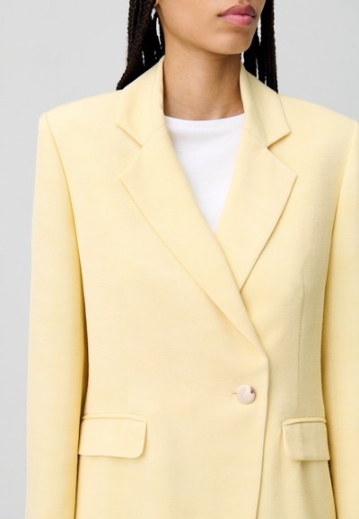 Blazer giallo doppiopetto con una texture liscia, revers a punta e un singolo bottone metallico. Include due tasche frontali.