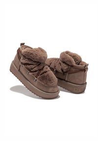 Botas marrones forradas con piel, de textura suave, punta redondeada y cordones elásticos. Presentan una suela robusta con un ligero relieve y una marca en el talón.