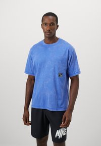Blue short-sleeve t-shirt with a textured surface, featuring a small black logo patch on the left chest. Paired with black shorts.