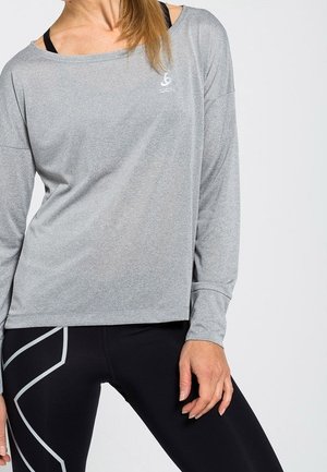 Kobieta ubrana w luźny szary top z długim rękawem na czarny sportowy stanik i czarne legginsy z szarymi geometrycznymi wzorami.