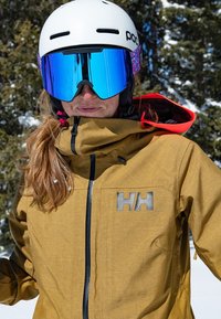 Chaqueta de invierno marrón texturizada con cremallera negra y logo reflectante 'HH'. Presenta detalles en rojo y un cuello alto, combinada con un casco blanco y gafas de sol azules.