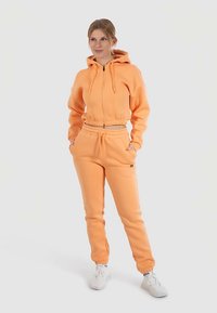 Sweat à capuche court orange avec poche kangourou et pantalon de jogging assorti. Le tissu est doux, avec une taille élastique et des poignets. Des baskets blanches complètent le look.