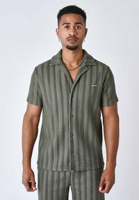 Camicia verde oliva a maniche corte con righe verticali, chiusura a bottoni sul davanti e una consistenza morbida. Presenta un colletto e un piccolo dettaglio con logo.