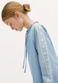 Hellblaue Bluse aus weichem Stoff mit Rüschen-Ausschnitt, Bindegürtel und dekorativer Stickerei entlang der Ärmel mit geometrischen Mustern.