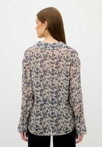 Camisa de manga larga en tejido semitransparente, con un fondo beige y estampado de flores en azul marino, cuello y puños con botones. Diseño texturizado y delicado.