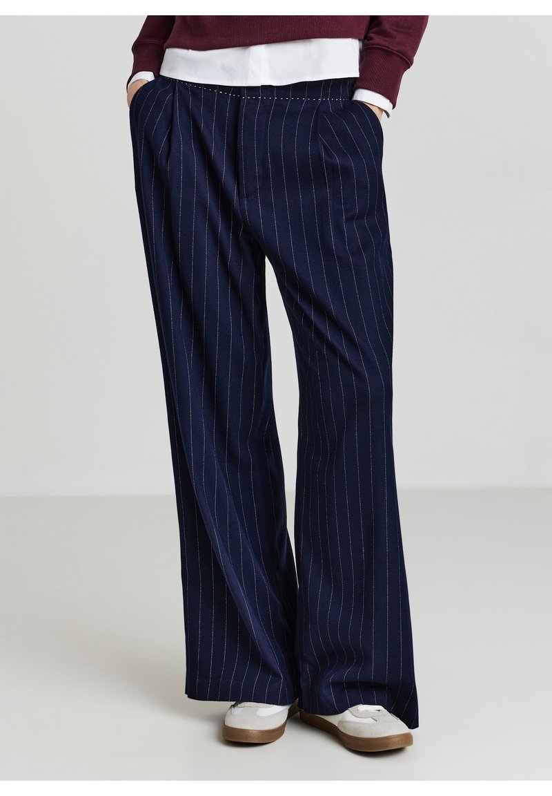 Personne portant un pantalon large bleu marine à fines rayures, les mains dans les poches et des baskets blanches avec des semelles en gomme.