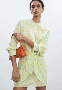Abito a quadri giallo e blu con dettaglio di nodo sulla parte anteriore. La modella tiene in mano una borsa arancione. Colletto tipo camicia, chiusura a bottoni e maniche lunghe.