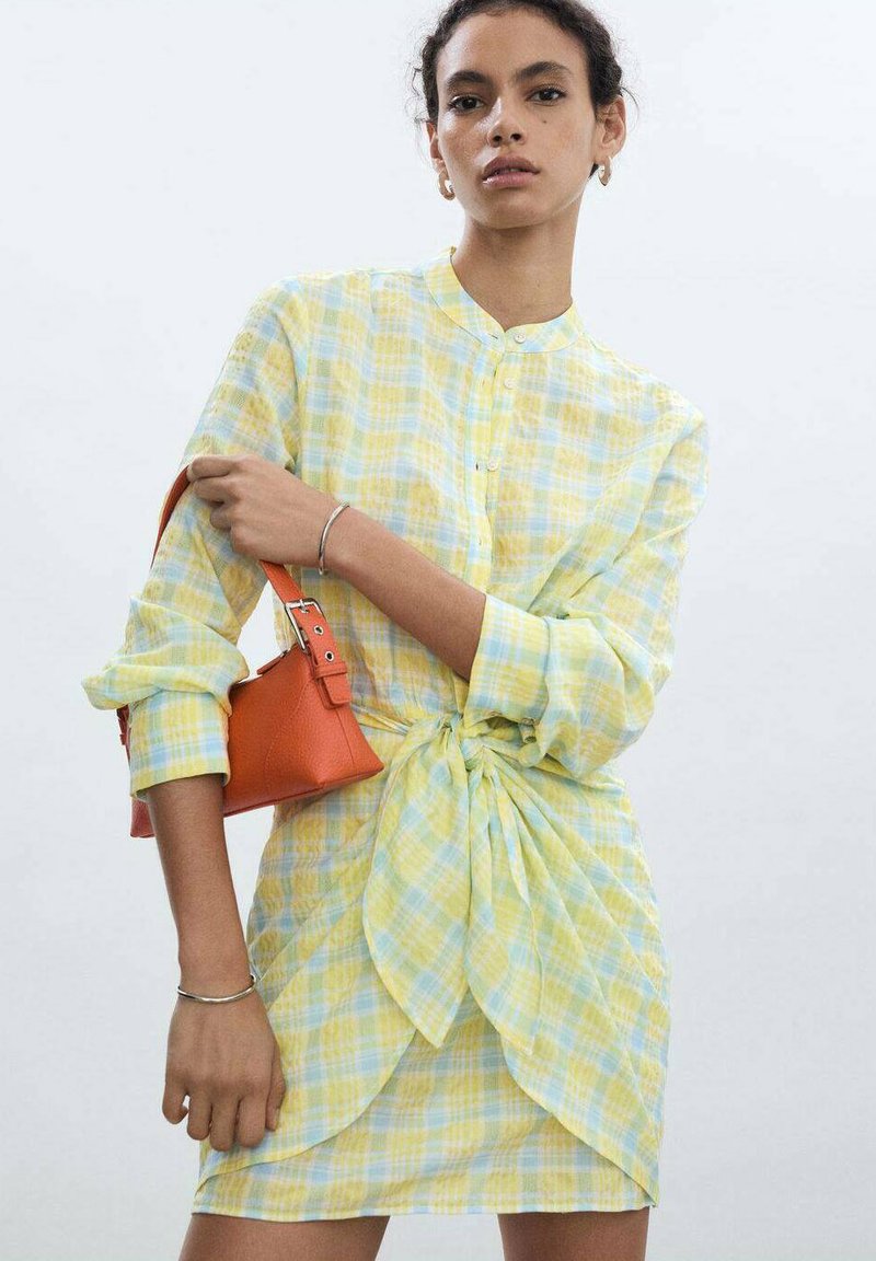 Abito a quadri giallo e blu con dettaglio di nodo sulla parte anteriore. La modella tiene in mano una borsa arancione. Colletto tipo camicia, chiusura a bottoni e maniche lunghe.