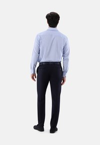 Mann in hellblauem Langarmshirt, schwarzem Gürtel, marineblauen Stoffhosen und schwarzen Schuhen, der mit dem Rücken vor einem weißen Hintergrund steht.