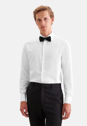 Jeune homme portant une chemise blanche avec un nœud papillon noir et un pantalon formel noir, debout devant un fond clair uni.