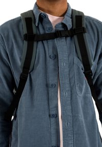Camisa de hombre de botones azul con una tela texturizada, que presenta un bolsillo en el pecho, usada bajo correas de mochila acolchadas grises aseguradas con cierres negros.
