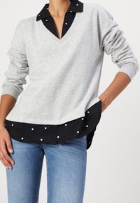 Felpa grigia con orlo e colletto a pois neri. Presenta un design a V e maniche lunghe leggermente a sbuffo.