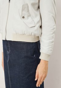 Lichtgrijze bomberjack met ribgebreide boorden en onderkant, voorzien van een ritssluiting en een schuine zak met zilverkleurige hardware, gecombineerd met een donkerdenim rok.