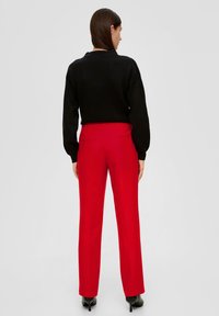 Czerwone spodni szyte na miarę z wysokim stanem, kieszeniami po bokach i kieszeniami z tyłu, w połączeniu z czarnym swetrem z prążkowanej tkaniny. Gładki materiał o wypolerowanej fakturze.