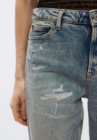 Jasnoniebieskie dżinsy z denimu o luźnym kroju, z przetarciami i postrzępionymi łatami, w standardowym fasonie z pięcioma kieszeniami i metalowymi dodatkami.