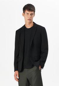 Sort blazer med stikkrave, to forlommer og en glat tekstur, parret med en sort ribbet t-shirt og grønne bukser.