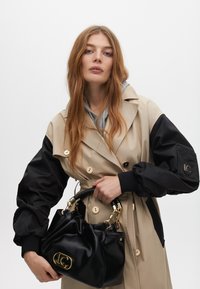 Beżowy płaszcz trench z czarnymi, bufiastymi rękawami, z złotymi guzikami i paskiem w talii. Trzyma czarną, teksturowaną torebkę z złotymi elementami.