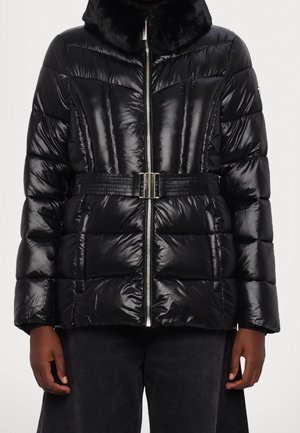 Doudoune noire brillante avec fermeture éclair, taille ceinturée et col haut avec bordure en fausse fourrure, portée par une personne à la peau foncée et portant un pantalon foncé.