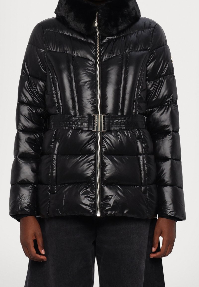 Doudoune noire brillante avec fermeture éclair, taille ceinturée et col haut avec bordure en fausse fourrure, portée par une personne à la peau foncée et portant un pantalon foncé.