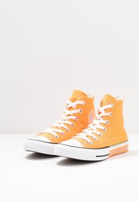 Pár vysokých oranžových plátěných tenisek se bílými tkaničkami a gumovými podrážkami, umístěných vedle sebe na bílé podložce.