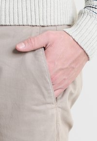 Beige bomull byxor i slim fit, med sido fickor och en subtil textur. En hand är placerad i höger ficka, delvis synlig.